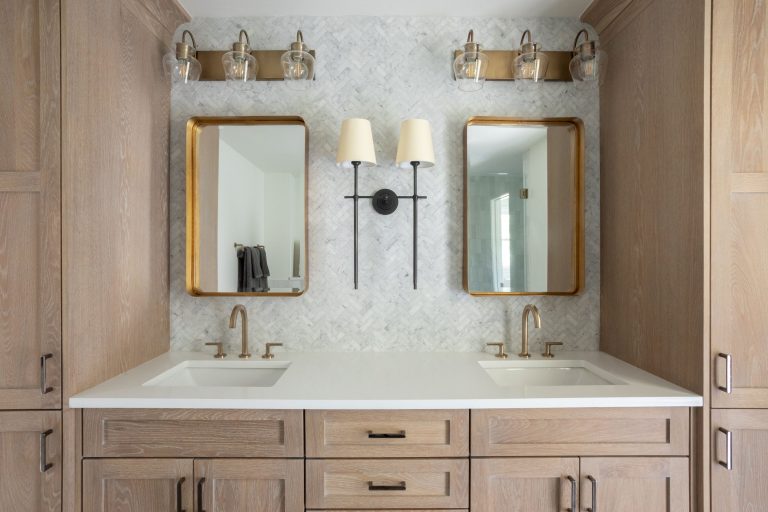 Vintage minimalist custom oak stained bathroom vanity with marble herringbone accent wall, featuring a double wallchiere and three-light wall sconces. Two gold-framed mirrors hang above matching sinks.