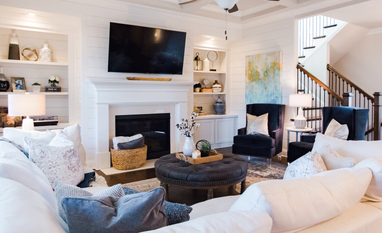 Main living room in bright white natural tones. White shiplap walls, white fireplace with tv hung above. Build in shelves on each side of the fireplace. White couch L couch with two navy accent chairs and round grey tufted ottoman coffee table.