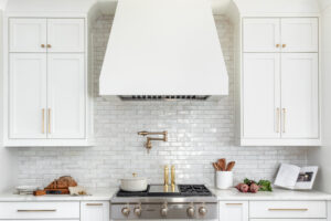 Modern white kitchen with gold hardware featuring stove and white range hood, cabinets, and subway tile.