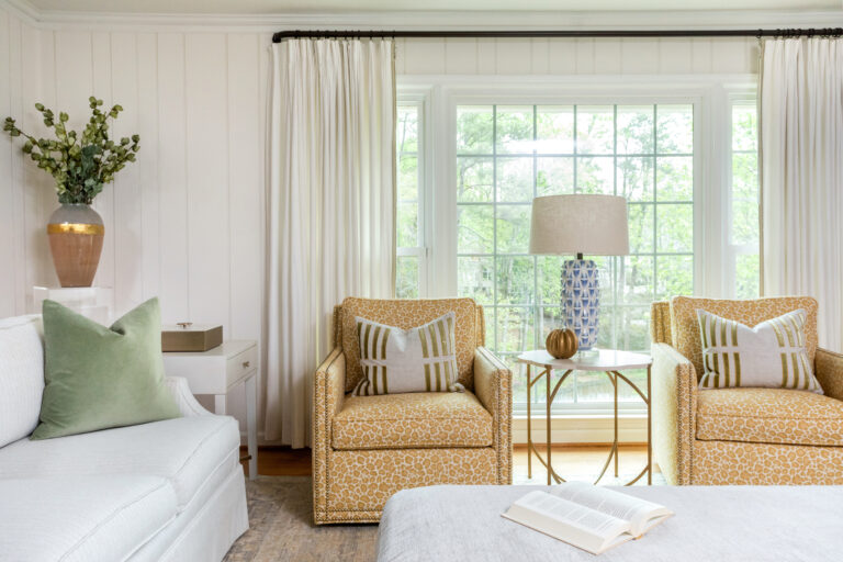 Modern feminine style family room featuring two yellow cheetah print accent chairs with gold nailhead trim. White marble top side table with gold legs with a patterned blue and white lamp and gold decor placed on top. White couch with green decorative pillow with a large rectangular ottoman. Black curtain rod hung against white shiplap wall with cream floor length curtains.