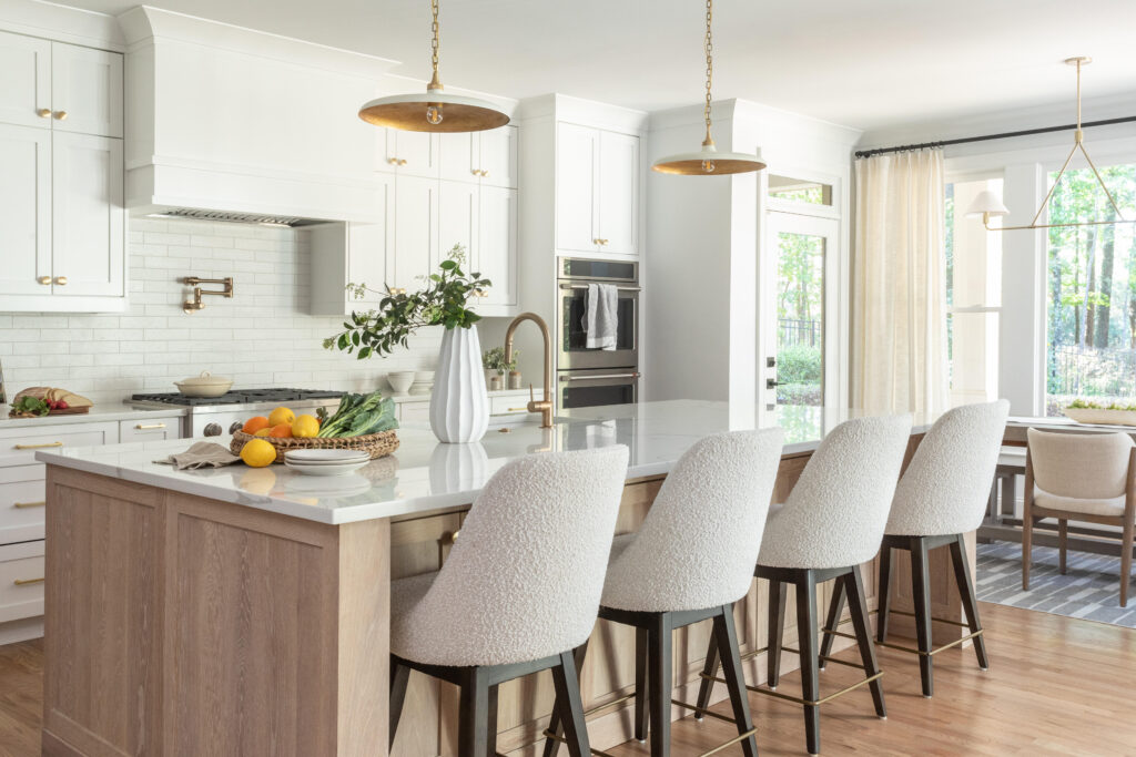 A newly renovated kitchen designed by Z & Co. Design Group, featuring a large wood island with marble countertop and white textured stools. Greenery in a white vase and fruit sit on the counter with white tile backsplash in the background. 