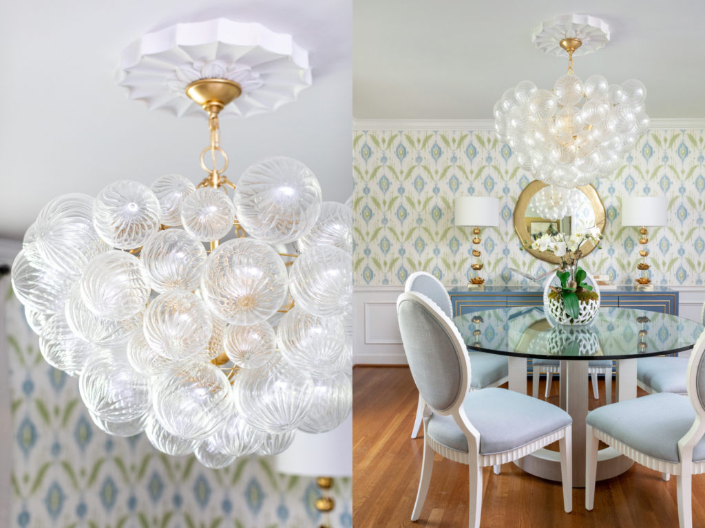 A bubble chandelier with a gold suspension system hangs above a round glass table with 4 light blue chairs around it. Gold lamps, a gold mirror, and blue and green patterned wallpaper make up the background. 
