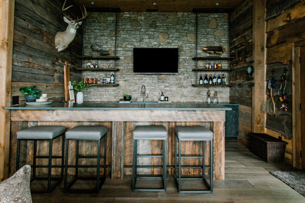 Wood cabin bar with four black and gray tools, decor mounted on the walls including a deer head, a black countertop, and a stone wall. 