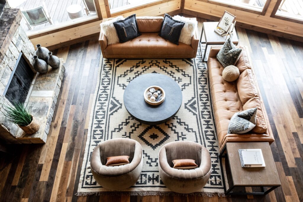 Mountain home living room with two-toned wood floors, a tribal black-and-white area carpet, 2 leather sofas, 2 upholstered chairs, and a round black coffee table. 