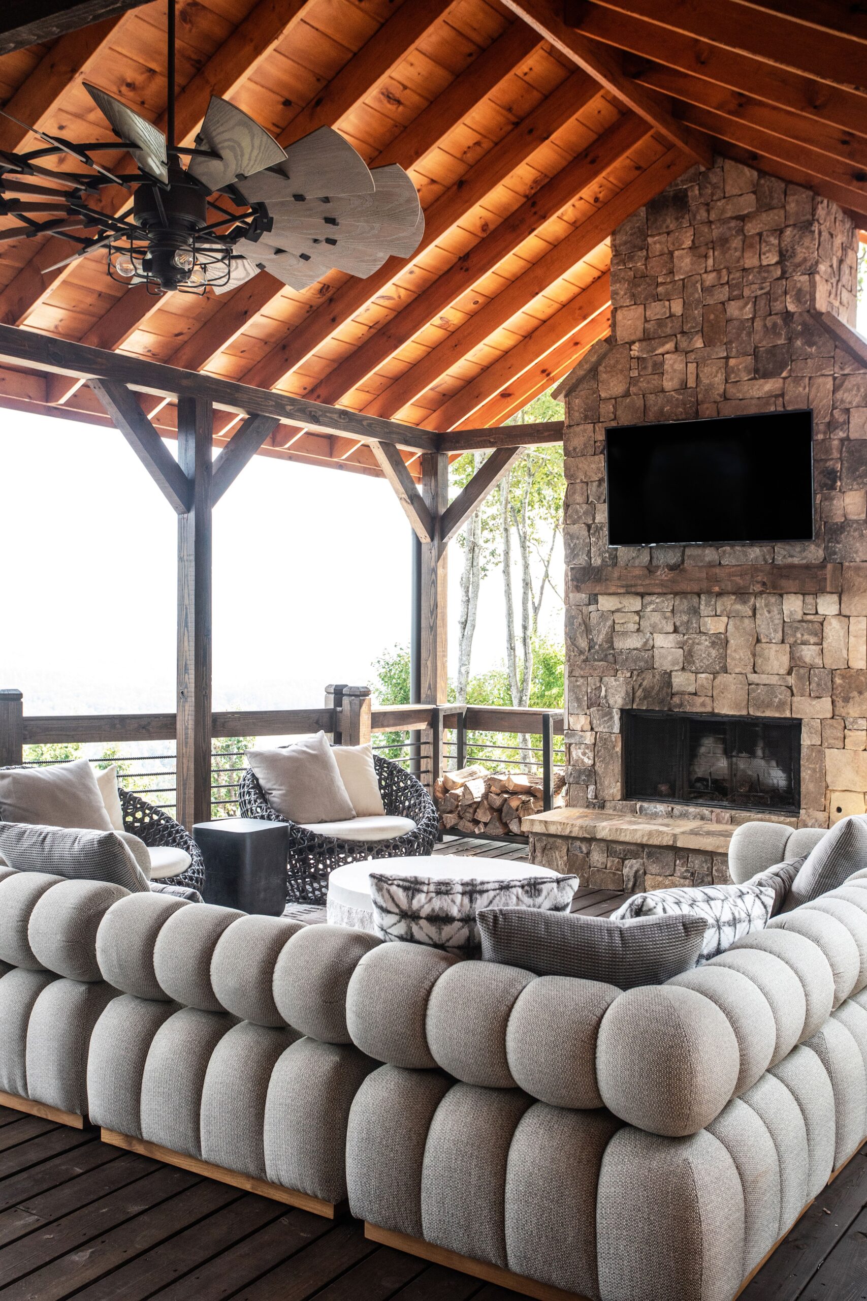 Outdoor balcony area with a gray sectional couch, Tv mounted on a stone fireplace, black metal and wood fan, with a cathedral roof. 