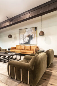A rust colored leather sofa sits under a large modern painting, with a brown steel beam running across the room and two bronze pendant lights hanging on either side of the sofa. Two olive-colored velour chairs sit in the foreground to the right of the sofa, with a black modern coffee table in the center.