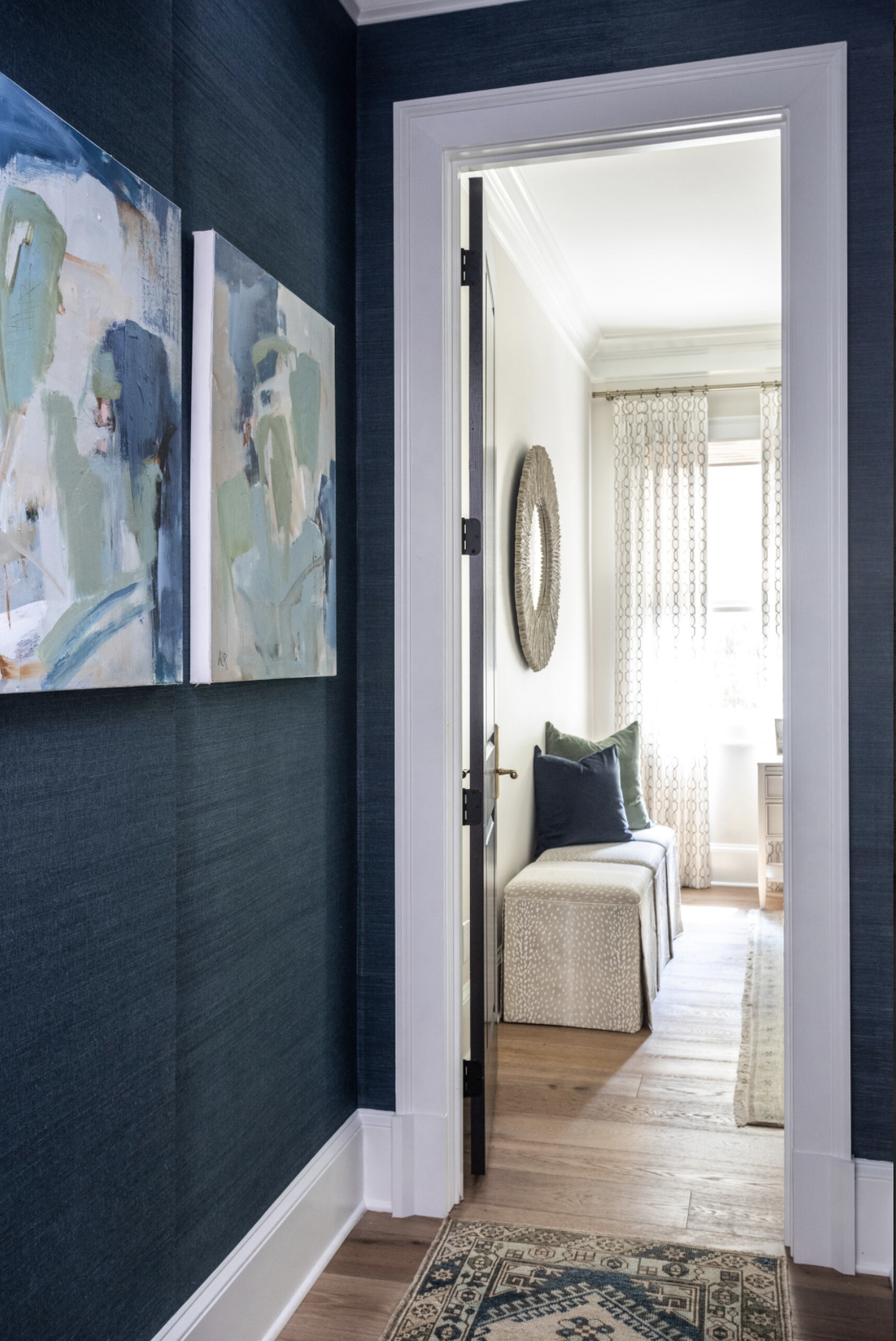 Dark blue grasscloth covers wall in hallway with white trim and two white, blue, and green abstract canvasses hanging on wall.