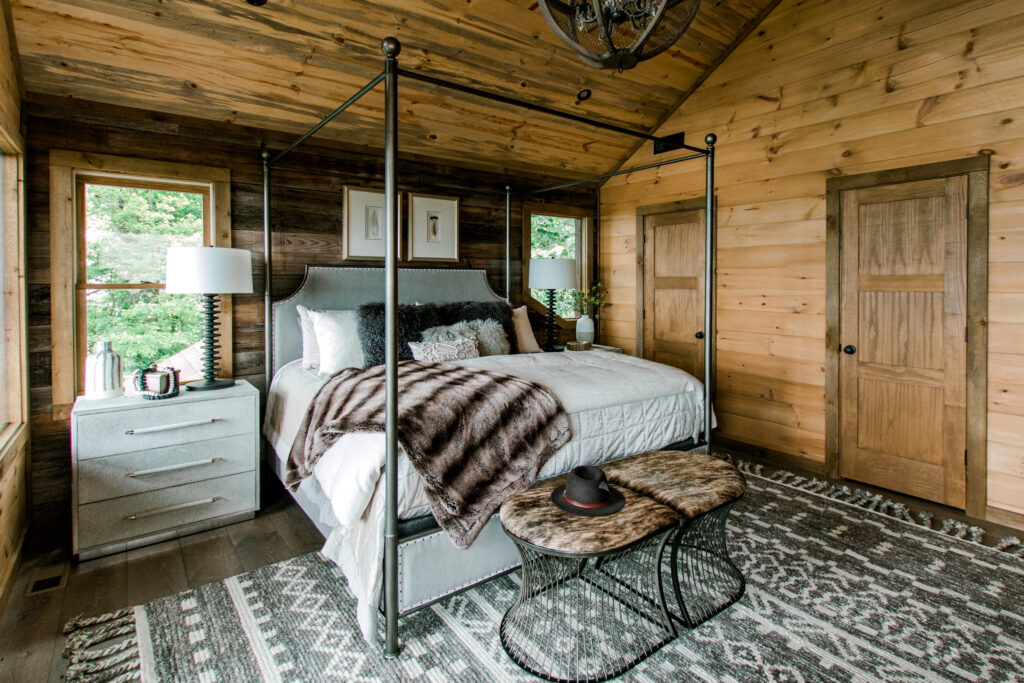 Cabin bedroom with fur throw, patterned rug, wood walls and flooring, fur bench, and other rustic decor pieces. 