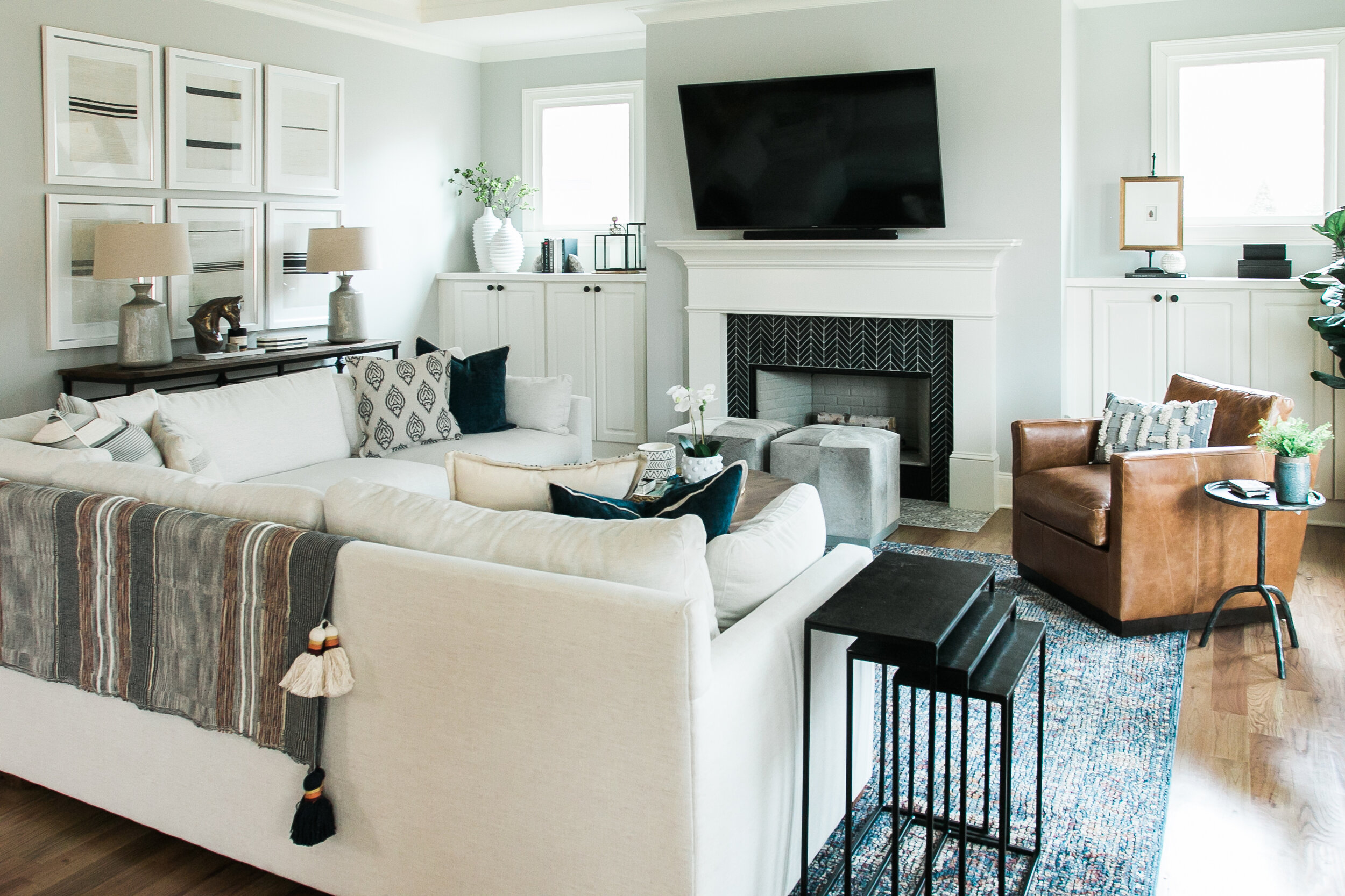 A cozy minimalist living room features white built-in cabinets on either side of a fireplace with white crown molding and a black herringbone tiled interior with a flatscreen on the mantle above it. A white couch and brown leather chair sit on a blue multicolored rug, with white trimmed windows above the cabinets on either side of the fireplace with natural light coming through. The walls are a light sea foam green with plants, lamps, and textured blankets adding warmth and dimension, with a set of black nesting tables sitting beside the couch in the foreground.