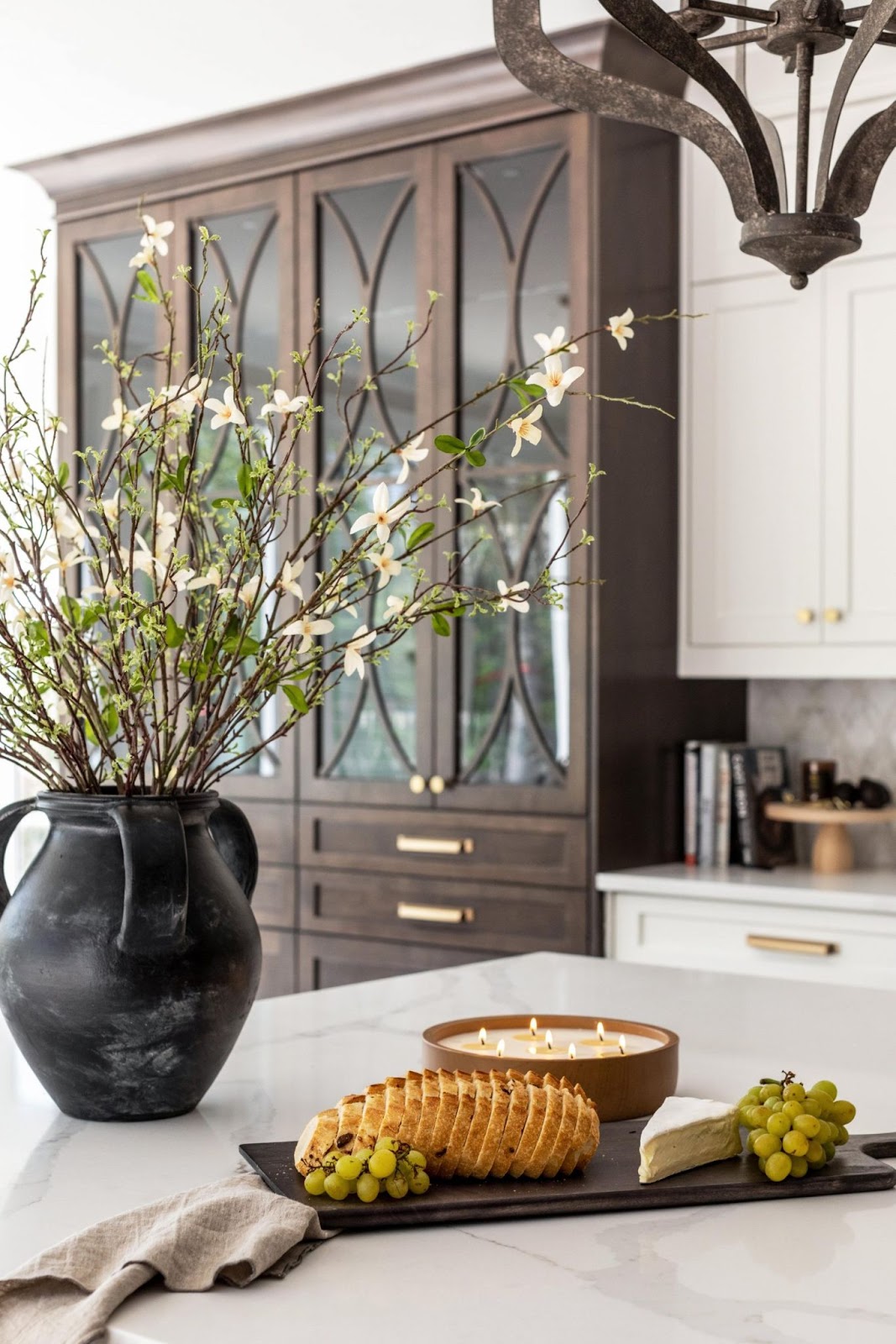 A Southern kitchen counter with a black serving tray that has a large candle, bread, grapes, and cheese on it. Branchy wildflowers pop out of black vase. 