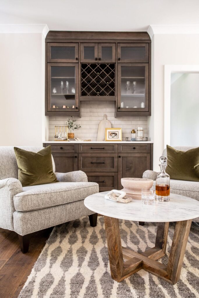 Bar area with built-in cabinets for glasses and wine bottle storage. Entertainment area with two gray chairs and round table with liquor decanter. 