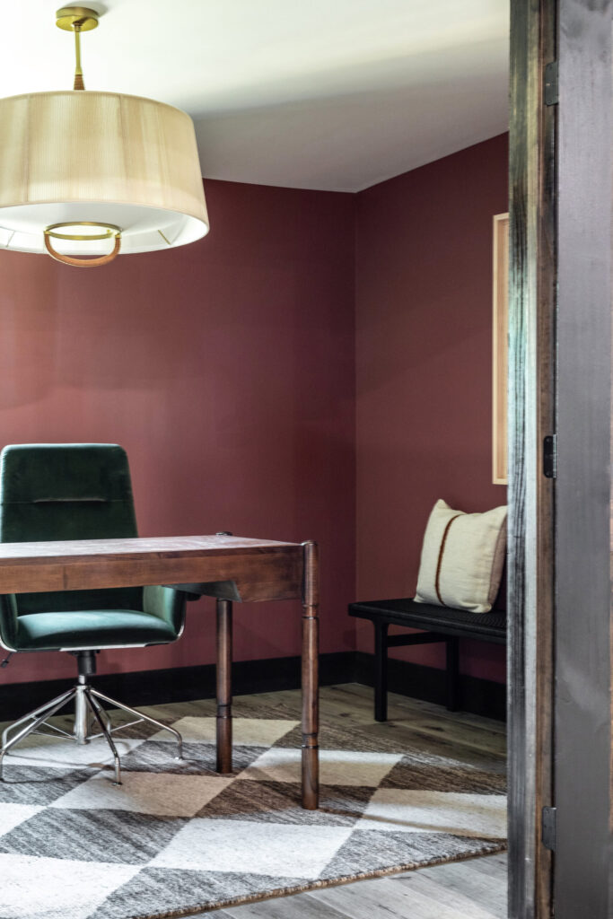 Office space with checked rug, emerald green chair, deep red walls, and wood desk. 
