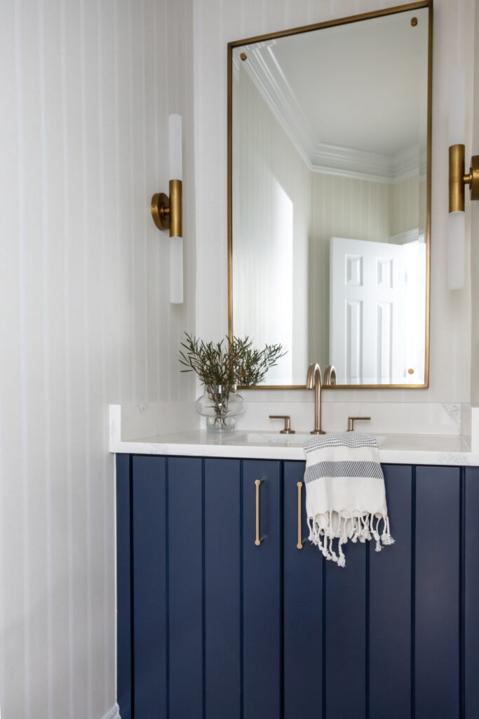 Bathroom with white shiplap walls, navy blue vanity, and brass framed mirror. 