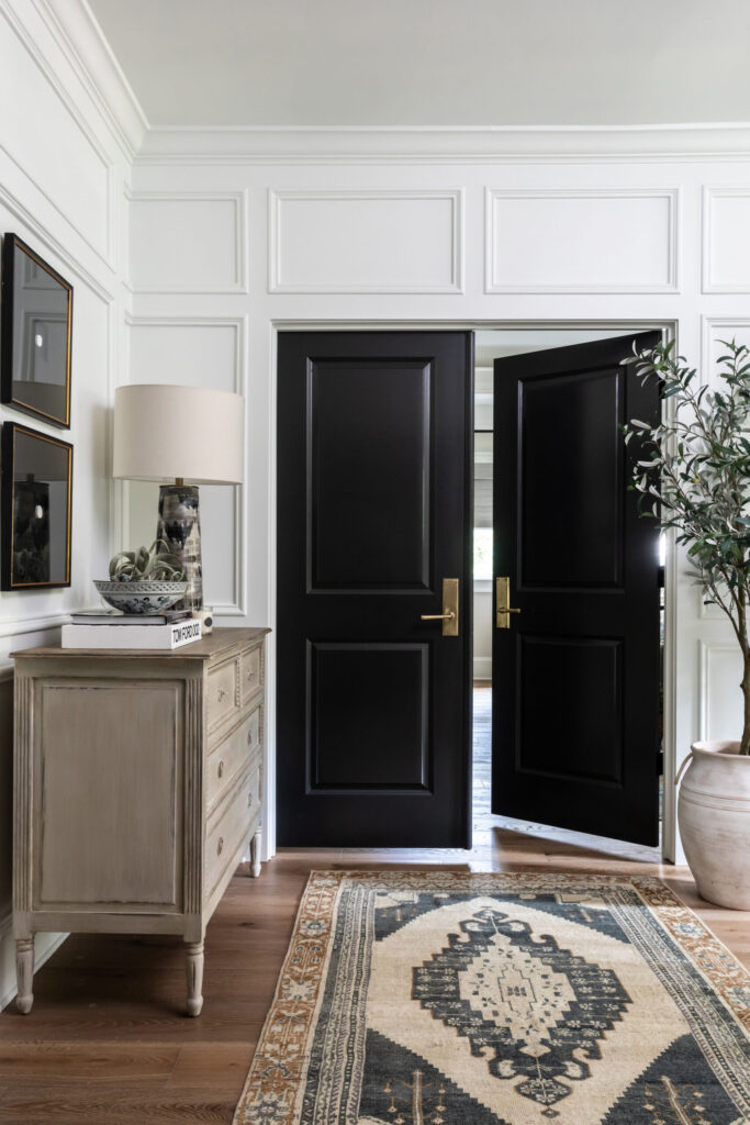 Black double doors, white wainscoating, patterned rug, beige sideboard. 