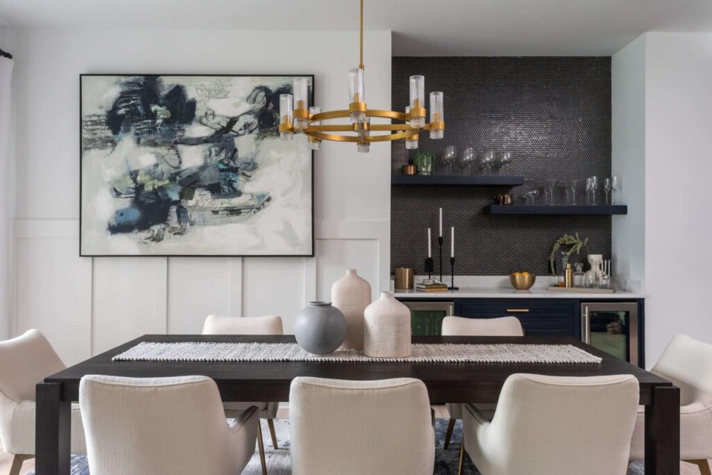 A stylish dining room with a large abstract painting on the wall, a dark wood dining table with eight white upholstered chairs, and a modern gold chandelier. The background features a wet bar area with dark cabinetry, a marble countertop, and shelves with glassware and decor.