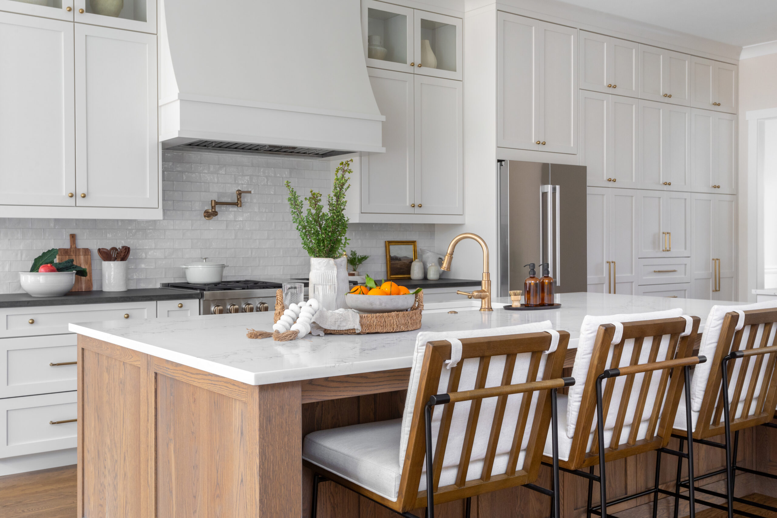 A modern cozy kitchen island with no overhead pendant lighting. In the background, a large white range hangs over a a chrome stove with a copper-piped faucet above it. White cabinets with copper knobs hang over a white subway tile backsplash with matching drawers underneath a black quartz countertop. The kitchen island and matching chairs have woodgrain bases with white cushions and a white quartz countertop featuring a copper faucet.