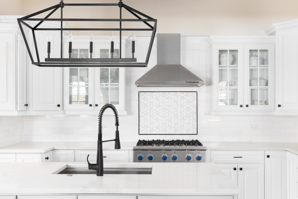 A geometric black metal frame supports a minimalist linear chandelier with four bulbs in a row over a white kitchen island with quartz countertops and a black faucet.