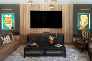 Contemporary living room with a dark feature wall, large flat-screen TV, brown leather sofa, matching armchair, and dark wood coffee table. Decor includes framed prints of hands holding drinks.