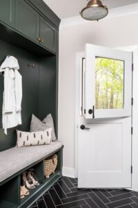 A cozy mudroom with dark green built-in storage, a cushioned bench, and black herringbone tile flooring, leading to a Dutch door that opens to a view of trees outside.