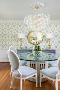 A dining room with bubble chandelier above round glass dining table and blue chairs.