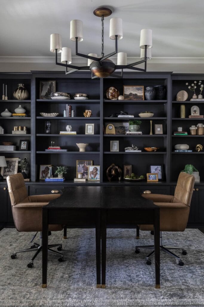Office space with two brown, leather chairs, lampshade chandelier, and navy bookshelf.