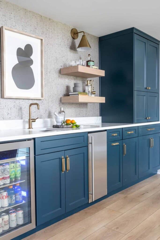 Wet bar with blue cabinets, cork wall covering, with mini fridge and muddling equipment.